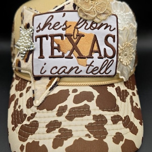 May include: A beige and brown trucker hat with a cow print design on the front and a patch that reads "she's from TEXAS i can tell." The hat is decorated with lace and a pearl-like embellishment.