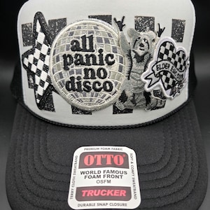 May include: Black and white trucker hat with a foam front. The hat features embroidered patches with the text "all panic no disco", a disco ball, a bear, and a checkered flag design. The hat has a durable snap closure.