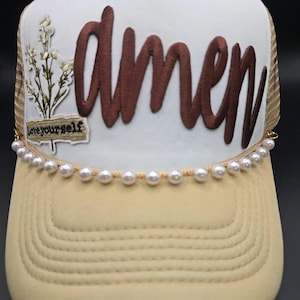 May include: A beige and white trucker hat with the word "amen" embroidered in brown script. The hat features a floral patch and the phrase "Love yourself." A string of pearls adorns the brim.