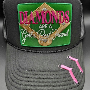 May include: Black trucker hat with a curved brim and mesh back. The front features a green patch with the phrase "DIAMONDS ARE A Girl's Best Friend" in pink script. Two pink earrings are pinned to the brim.