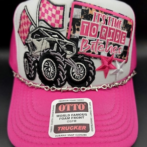 May include: A pink and white trucker hat with a mesh back. The front features a monster truck graphic, checkered flags, and the text "IT'S TIME TO RIDE Bitches". A barbed wire design accents the brim.