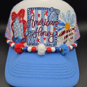 May include: A white and blue baseball cap with the text "American Honey" embroidered on the front. The cap features red, white, and blue accents, including a beaded band with pom-poms and a floral design, and a blue brim.