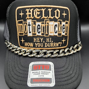 May include: A black trucker hat featuring a rectangular patch with gold embroidery that reads HELLO motherfucker HEY, HI, HOW YOU DURRN?. The hat includes a leopard print chain across the base of the patch and an Otto brand sticker on the brim.