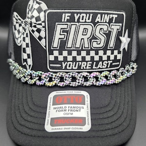 May include: A black trucker hat featuring a rectangular patch with the white embroidered text IF YOU AIN'T FIRST YOU'RE LAST, a checkered bow, a small star, and a metallic iridescent chain across the brim. A sticker on the brim reads OTTO.