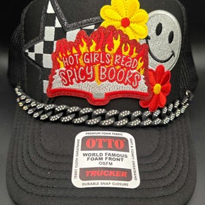 May include: Black trucker hat with a mesh back, featuring a chain detail and patches. Patches include a checkered star, a smiley face, and a design that reads "HOT GIRLS READ SPICY BOOKS".