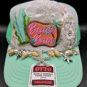 May include: A mint green trucker hat with a white front panel, embellished with a "Beach Babe" patch, seashells, pearls, and starfish charms. The hat features a mesh back and a snap closure. The hat has the text "OTTO" and "TRUCKER" on the tag.