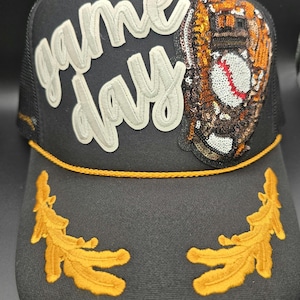May include: Black baseball cap with a white "game day" patch and a sequined baseball glove patch. The cap has a gold embroidered design on the brim.