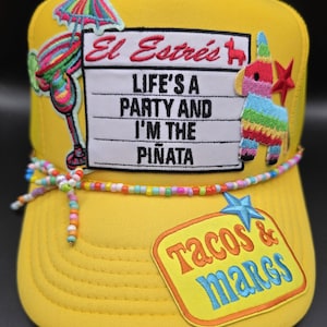 May include: A bright yellow trucker hat featuring a central patch with the text "El Estrés LIFE'S A PARTY AND I'M THE PIÑATA". It includes embroidered patches of a cocktail, a piñata, and a star, plus a colorful beaded string across the brim.