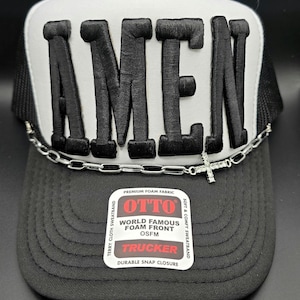 May include: Black and white trucker hat with the word "AMEN" embroidered in bold, black letters. A silver chain with a cross charm is draped across the brim. The hat features a "OTTO" label.