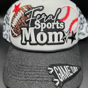 May include: A white trucker hat with a dark gray brim featuring a sequined football, a baseball graphic, and the text Feral Sports Mom. A black pennant patch with the text GAME DAY is on the brim, accented by red and black star appliqués.