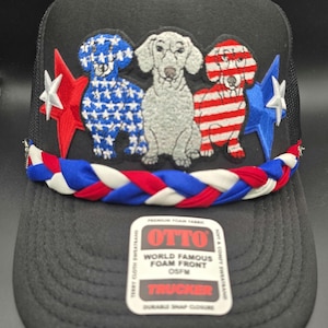 May include: Black trucker hat with a mesh back and a foam front. The front features three dog designs with patriotic patterns, stars, and stripes. A braided red, white, and blue band accents the brim. The hat includes the text "OTTO" and "TRUCKER".