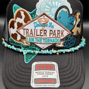 May include: A black trucker hat featuring a central diamond-shaped patch with the text "You are the TRAILER PARK I AM THE TORNADO". The hat is decorated with various patches including cow print and turquoise sequins, and a turquoise bead trim.