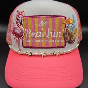 May include: A white and pink trucker hat with a neon pink brim. The hat features a flamingo, a beach scene with the word "Beachin'", and a coconut drink. The brim is decorated with pearls and starfish.