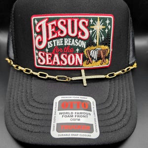 May include: A black trucker hat featuring a rectangular patch with the text Jesus is the reason for the season and a manger scene. A gold-toned chain with a cross charm is attached across the front. A sticker on the brim reads Otto.