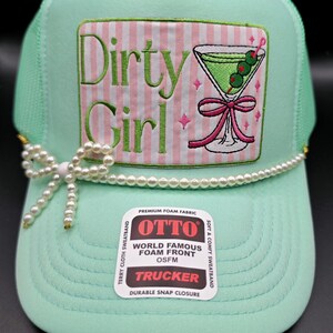 May include: A mint green trucker hat featuring a rectangular patch with pink and white stripes, the text "Dirty Girl" in green, and an embroidered martini glass with a pink bow. A string of white faux pearls with a bow accent sits above the brim.