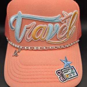 May include: A coral-colored trucker hat with the word "Travel" embroidered in blue, yellow, and pink. A silver chain with pearl accents and a small airplane charm adorns the brim. A patch with a blue star and the words "Adventure Awaits" is on the front.