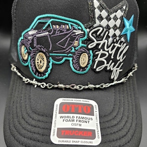 May include: Black trucker hat with a mesh back and a curved brim. The front features an embroidered off-road vehicle design with the text "Shitt Buff". A barbed wire design accents the brim.
