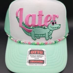 May include: A mint green and white trucker hat with the word "Later" in pink script above a cartoon alligator wearing sunglasses. The hat has a beaded band and a "OTTO" label.