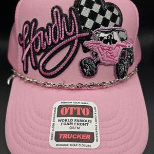 May include: Pink trucker hat with a mesh back and a foam front. The hat features the word "Handy" in cursive, a pink off-road vehicle, and a checkered flag. A barbed wire design accents the brim. The hat has a "TRUCKER" label.