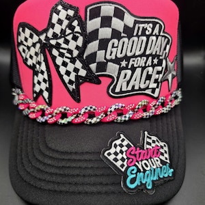 May include: A hot pink and black trucker hat featuring a checkered bow patch, a checkered flag patch with the text IT'S A GOOD DAY FOR A RACE, a silver star, a decorative chain across the brim, and a patch on the brim reading Start YOUR Engines.