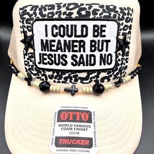 May include: A cream-colored trucker hat featuring a black and white leopard print patch with the embroidered text I COULD BE MEANER BUT JESUS SAID NO. The hat is adorned with black star accents and a beaded trim with black and tan beads and a cross.