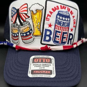 May include: A navy blue trucker hat with a white front panel featuring patches of beer cans, a beer glass, and a patriotic bow. The hat has the text "It's a bad day to be a cold American Beer."