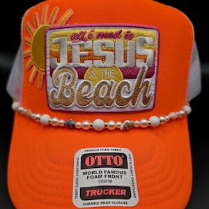 May include: A bright orange trucker hat with a white mesh back and a foam front. An embroidered patch features a yellow sun and the text "all i need is JESUS & THE Beach". A string of white beads and small seashells accents the base of the crown.