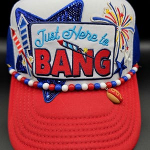 May include: A patriotic trucker hat with a red brim and white crown. The hat features a beaded necklace and a charm of a hot dog. The text "Just Here to BANG" is embroidered on the front, along with firework and star designs.