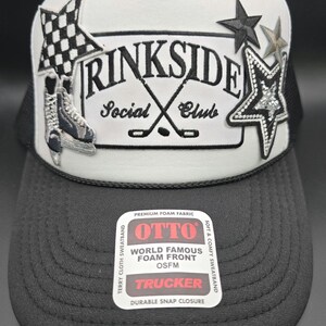 May include: A black and white trucker hat with the text "Rinkside Social Club" and decorative stars and a checkered flag. The hat has a black brim and mesh back, and a white front panel. The hat has a "OTTO" tag.