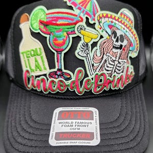 May include: Black trucker hat with a mesh back and a foam front. The front features embroidered images of cocktails, a skeleton in a sombrero, and the text "Cinco de Drinks". The hat also has the text "TEQUILA" on it.