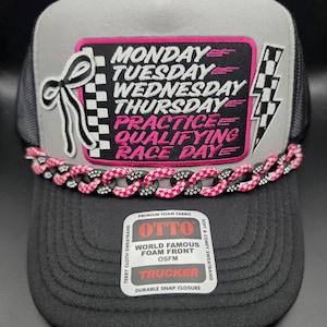 May include: A gray and black trucker hat featuring a rectangular patch with the text MONDAY TUESDAY WEDNESDAY THURSDAY PRACTICE QUALIFYING RACE DAY, a black bow, a checkered lightning bolt, and a pink and black chain trim across the brim.