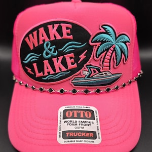 May include: A bright pink trucker hat featuring a black oval patch with the embroidered text WAKE & LAKE, a teal palm tree, and a motorboat. A silver-toned chain with black rhinestones accents the base of the crown, and an Otto brand sticker is attached.