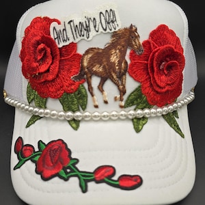 May include: White trucker hat with a horse graphic, red rose embroidery, and the text "And They're Off!". The hat features a pearl beaded trim and a mesh back.