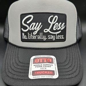 May include: A black and gray trucker hat with the text "Say Less" embroidered on a black patch. The hat has a gray front panel and a black brim. The hat also has a white tag that says "OTTO".