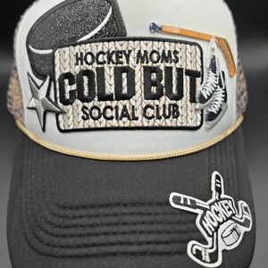 May include: A trucker hat with a white top and black brim. The hat features a hockey puck, star, and the text "HOCKEY MOMS COLD BUT SOCIAL CLUB." Additional hockey-themed patches are also visible.