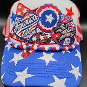 May include: A white and blue trucker hat featuring a red sequined star, a heart-shaped patch reading America 250th Birthday, a circular star patch, and a raccoon graphic holding a flag. The brim has white stars, and a red, white, and blue bead trim.