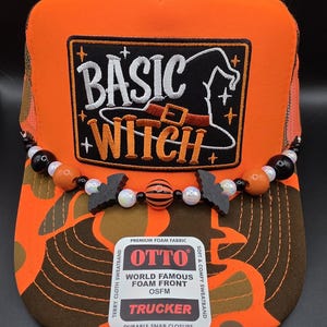 May include: An orange trucker hat with a black patch featuring the text BASIC WITCH and a witch hat graphic. The brim has an orange and brown camouflage pattern. A beaded string with black bat charms and orange, black, and white beads accents the front.