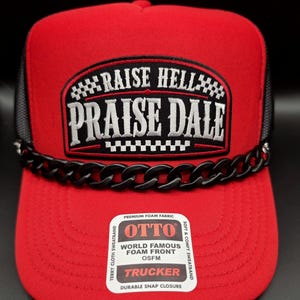 May include: A red trucker hat with a black mesh back and a black plastic chain across the base of the crown. The front features a black patch with white text reading RAISE HELL PRAISE DALE. A white sticker on the brim reads OTTO TRUCKER OSFM.