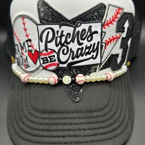 May include: A black and white baseball cap with the text "Pitches Be Crazy". The cap features baseball-themed embellishments, including baseballs, a heart, and the number 3. A pearl and baseball bead chain adorns the brim.