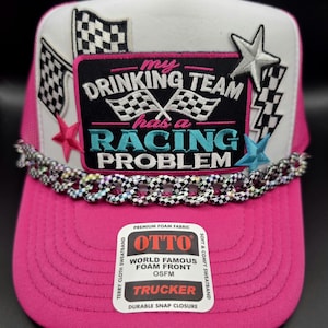 May include: A bright pink and white trucker hat featuring a black rectangular patch with the text "my DRINKING TEAM has a RACING PROBLEM" and checkered flags. The hat is decorated with pink, silver, and blue stars and a checkered pattern chain.