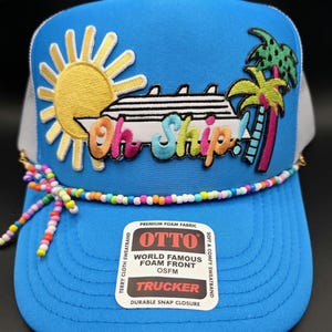 May include: A blue trucker hat with a white mesh back and a beaded accent. The front features a colorful embroidered design of a cruise ship, sun, and palm trees, with the text "Oh Ship!". The hat has a snap closure.