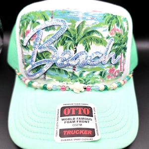 May include: A mint green trucker hat featuring a white front panel with a tropical palm tree print and the word Beach in blue sequins. A beaded strand with a small white turtle charm sits above the brim. An Otto brand sticker is on the brim.