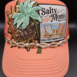 May include: Coral-colored trucker hat with a mesh back and a curved brim. The front features a patch with a palm tree, a sequined sun, and the text "Salty Moms Beach Club." A beaded shell and pearl accent adorns the brim.