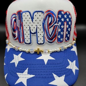 May include: A white trucker hat with the word "AMEN" in red, white, and blue lettering. The brim is blue with white stars. A beaded strand with a gold cross accents the hat.