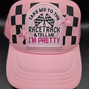 May include: A light pink trucker hat featuring a black and white checkered pattern on the sides. A central patch displays two checkered flags and the text "TAKE ME TO THE RACETRACK & TELL ME I'M PRETTY" in black and pink, accented by two pink stars.