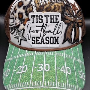 May include: A white trucker hat with a brown mesh back and a green glitter brim designed like a football field with yard lines. The front features a patch reading TIS THE football SEASON, a leopard print football helmet, a football, and a bow.