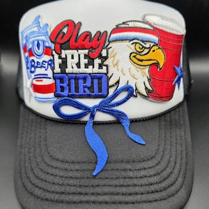 May include: A white and black trucker hat featuring embroidered patches of a beer can, a red party cup, and a bald eagle wearing a headband. The text Play FREE BIRD is embroidered in red, white, and blue, with a blue ribbon accent on the front panel.