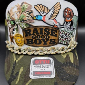 May include: A white and camouflage trucker hat featuring a brown leather patch with the text RAISE GOOD BOYS, embroidered patches of a flying duck, a dog in a crate, and antlers, plus a camouflage chain accent with rhinestones.