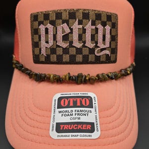 May include: A coral-colored trucker hat with a brown and black checkered patch that reads "petty" in glittery pink letters. The hat features a beaded band and a "OTTO" label.
