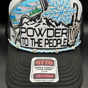May include: A black and white trucker hat with the text "POWDER TO THE PEOPLE." The hat features a ski goggles graphic, a snowflake, and a beaded band. The hat has a black brim and a white front panel.